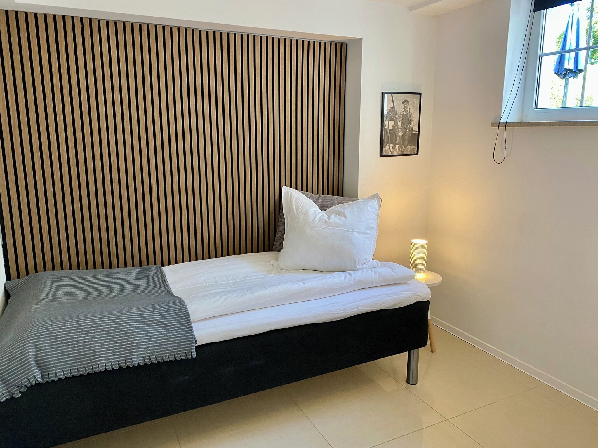 A modern bedroom with a black bed, white bedding, a wooden slat headboard, a bedside lamp, and a framed picture on the wall.