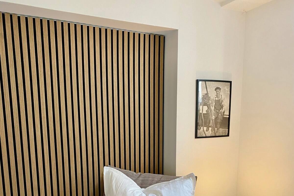 Una cama bien tendida con sábanas blancas y cabecera negra, una mesita de noche con lámpara, y una fotografía enmarcada en la pared.