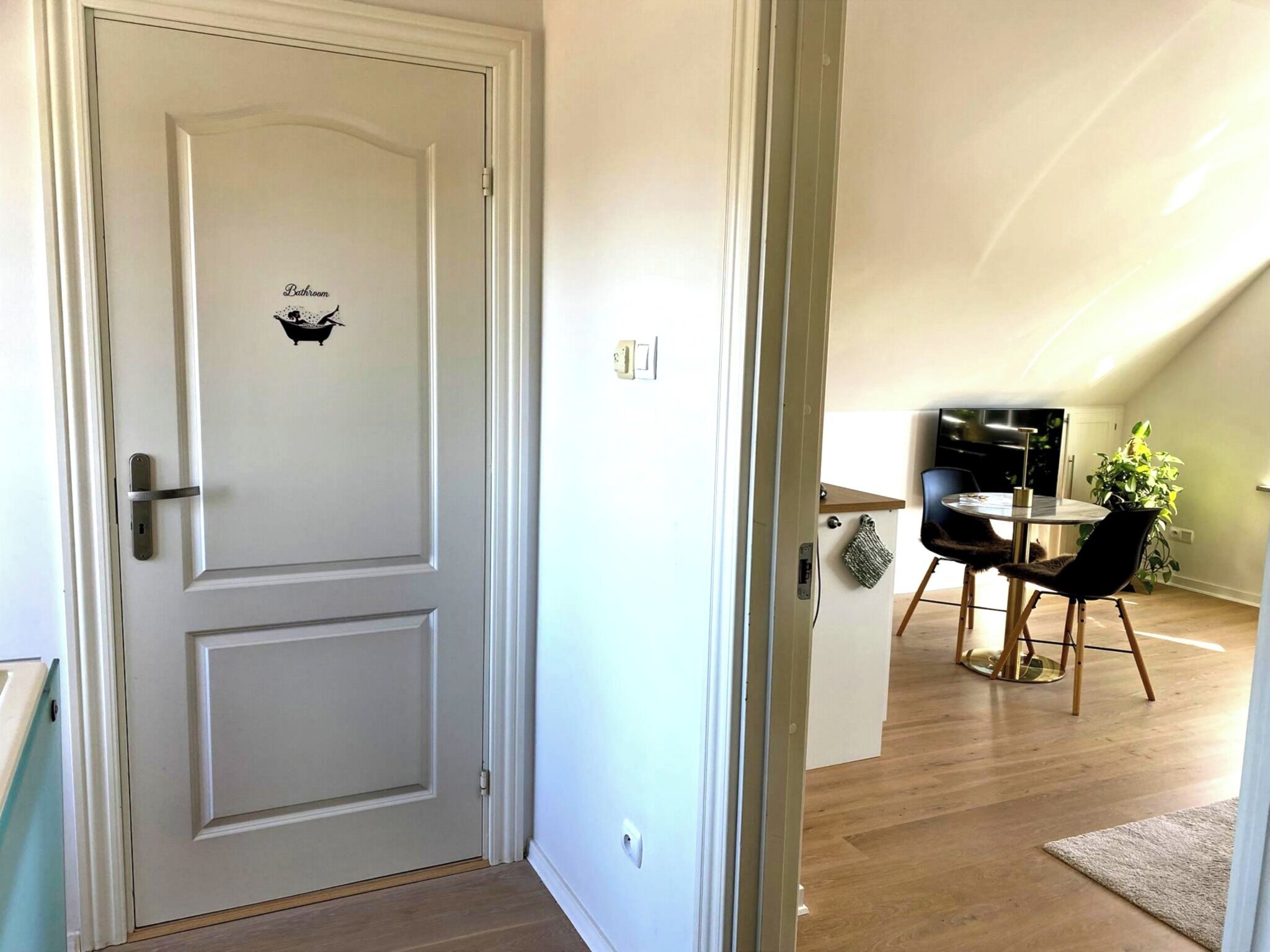 Una porta bianca con il segnale di bagno, che dà accesso a una sala da pranzo con un tavolo rotondo e sedie, e una pianta.