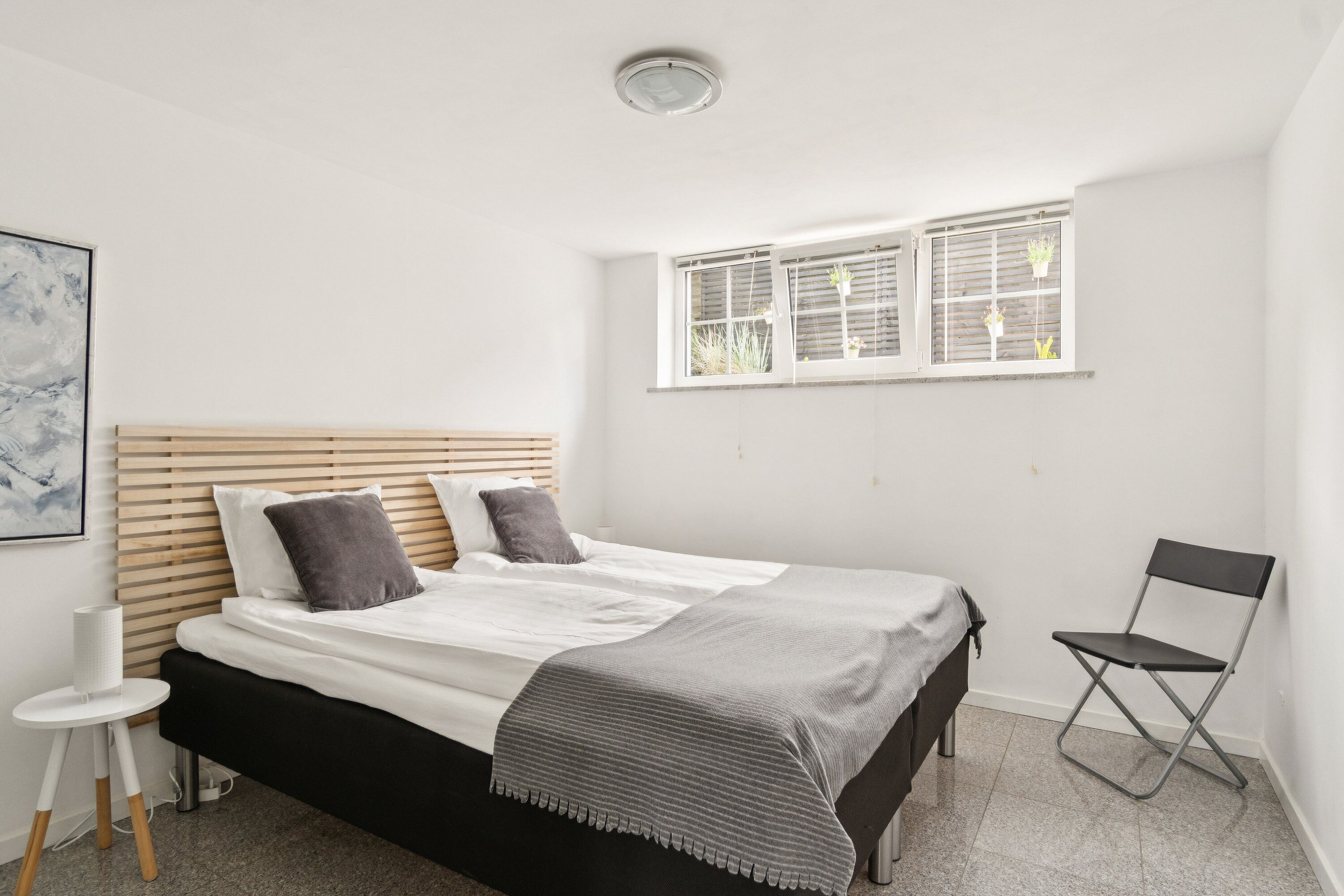 Une chambre à coucher avec un lit, une fenêtre avec des stores, une chaise et une petite table.