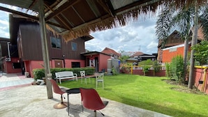 A covered outdoor seating area with a table and chairs, a grassy lawn, and a building with a red facade in the background. - Ho Hostel (Chiang Mai)