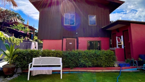 A two-story house with a red and dark brown exterior, a small front porch, and a white bench in the front yard. - Ho Hostel (Chiang Mai)