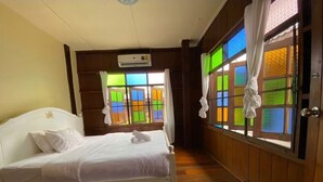A bedroom with a bed, wooden flooring, a window with colorful glass panes, and an air conditioning unit. - Ho Hostel (Chiang Mai)