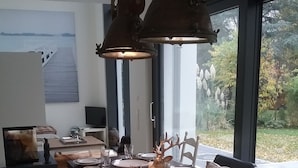 A dining area with a wooden table set for four, surrounded by chairs. A large window offers a view of greenery outside. - Rethorn Haus am See (Ganderkesee)