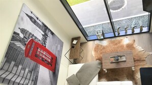 A modern living room with a large wall-mounted artwork of a cityscape, a white sofa with cushions, a wooden coffee table, and a glass window with a view of a city street and a building. - Rethorn Haus am See (Ganderkesee)