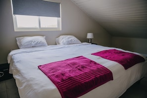Uma cama de solteiro com roupa de cama branca e duas mantas rosa vibrante, uma luminária de cabeceira e uma janela com persianas.