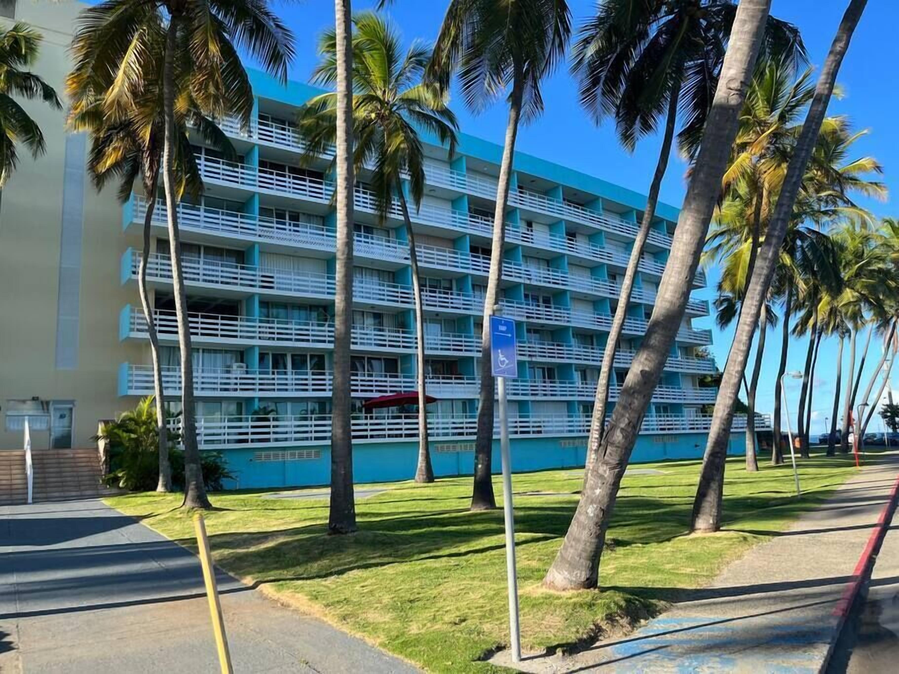 A multi-story hotel with a blue facade, white balconies, and palm trees in the foreground.