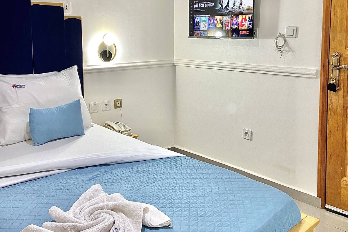 Hotellihuone, jossa on sänky, seinälle asennettu televisio, yöpöytä ja ovi.