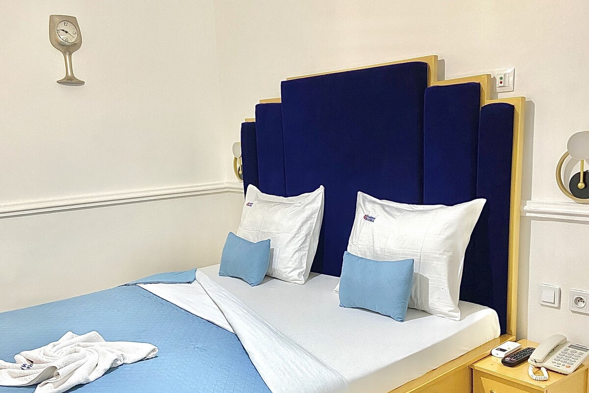 A neatly made bed with blue and white bedding, a bedside table with a phone and a lamp, and a wall clock.