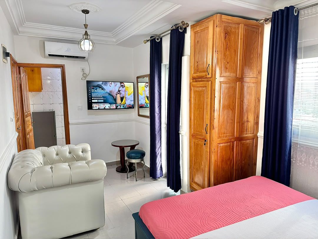 Habitación de hotel con cama, zona de estar, armario, televisión y ventana con cortinas.