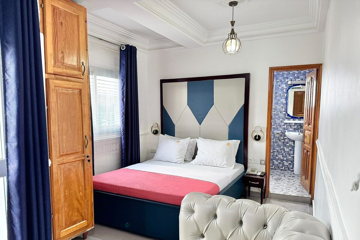 Een slaapkamer met een bed, een kledingkast, een nachtkastje en een badkamer zichtbaar door een geopende deur.