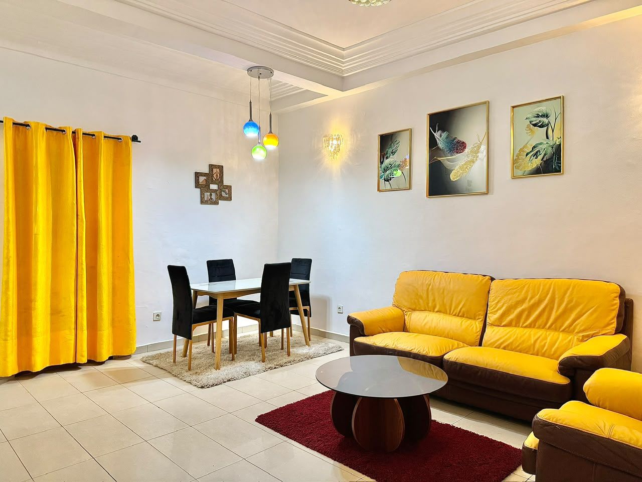 Una sala de estar con un sofá amarillo, una mesa de comedor con sillas, una araña y obras de arte en la pared.