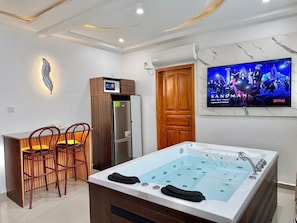 A modern bathroom with a large bathtub, wooden chairs, a built-in TV, and a wall-mounted air conditioning unit. - Residence La Marina (Douala)