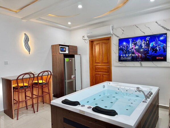 A modern bathroom with a large bathtub, wooden chairs, a built-in TV, and a wall-mounted air conditioning unit. - Residence La Marina (Douala)