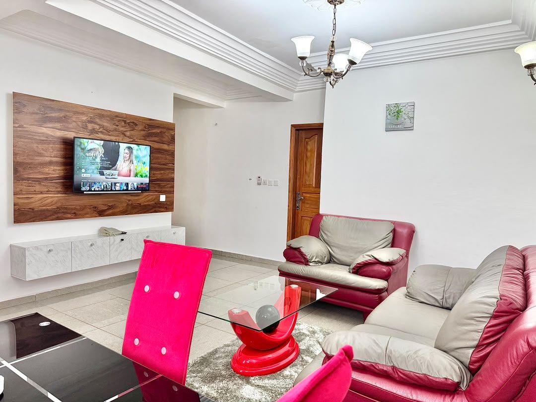 Una sala de estar moderna con un televisor de pantalla plana montado en un panel de madera, un sillón rojo, un sofá color beige y una mesa de centro de vidrio.
