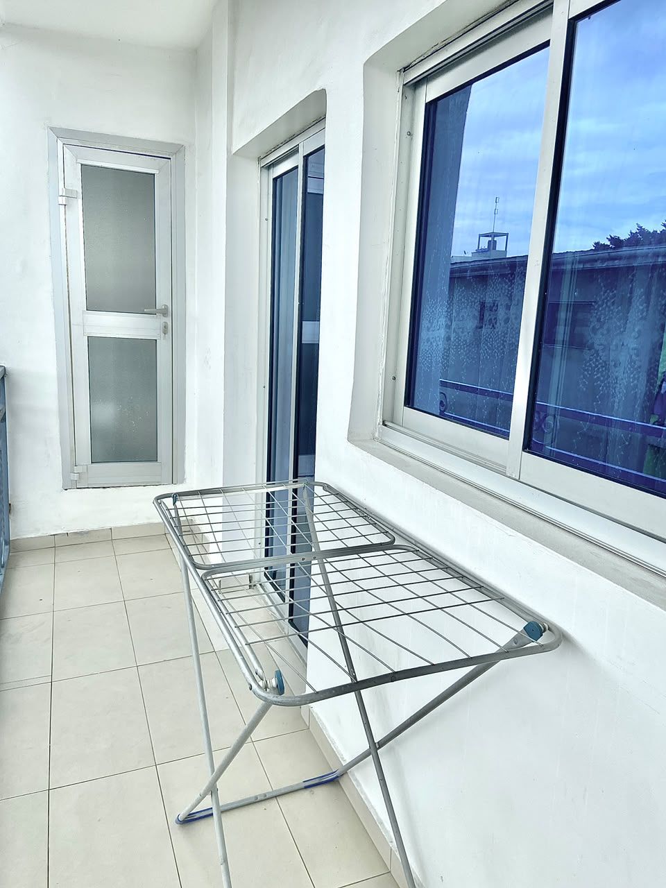 Un balcón con un tendedero metálico y una ventana con vistas a edificios.