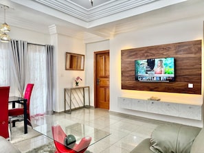 Een moderne woonkamer met een flatscreen televisie aan een houten wand, een glazen salontafel, rode en grijze stoelen en een houten deur.