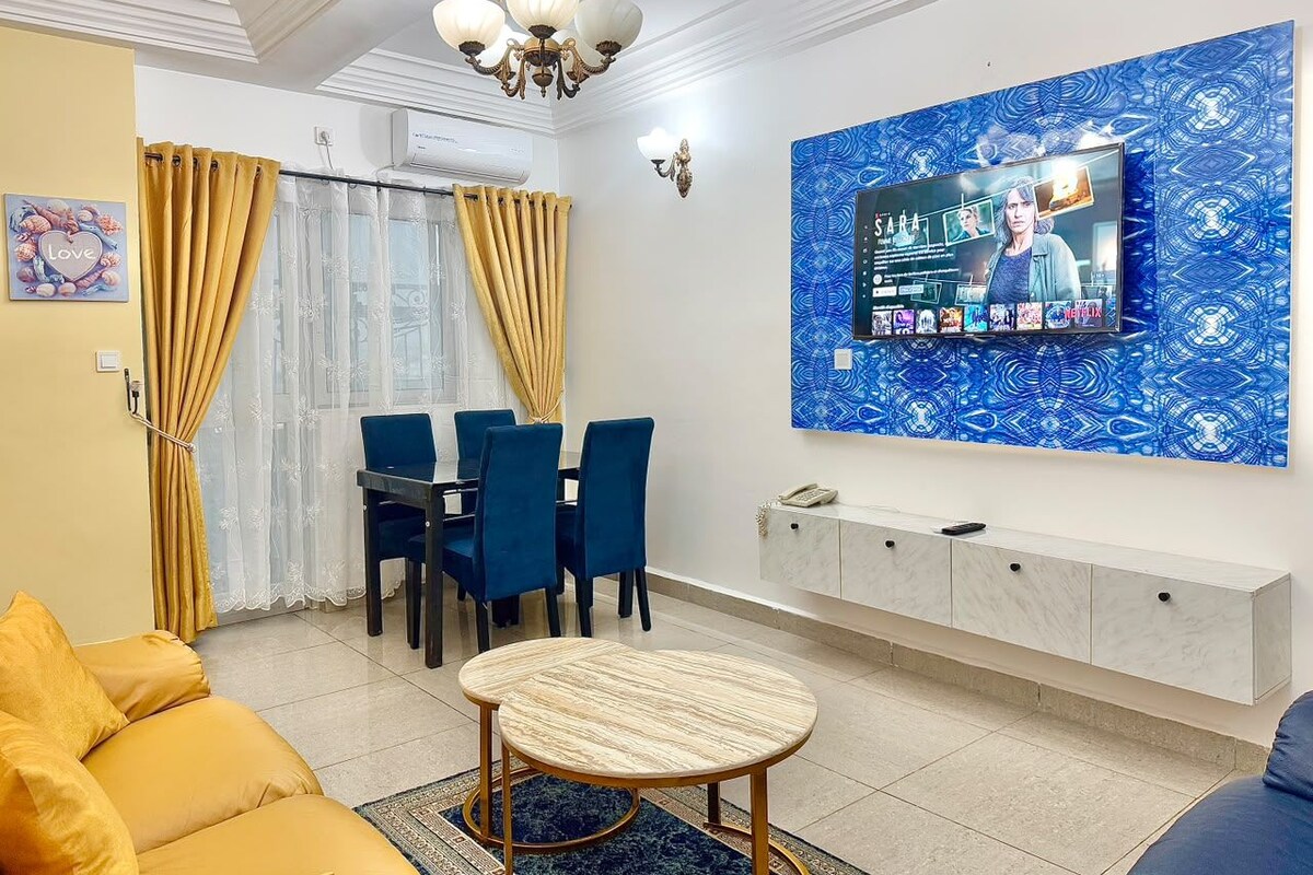 Een woonkamer met een bank, een eettafel met stoelen, een aan de muur bevestigde televisie en een kunstwerk met een patroon erboven.