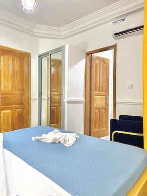 A hotel room with a bed, a wooden wardrobe, a mirror, and a door leading to another room. - Residence La Marina (Douala)