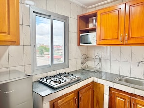 A kitchen with a gas stove, wooden cabinets, a microwave, and a window with a view of buildings. - Residence La Marina (Douala)