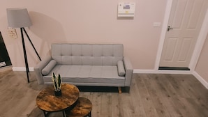A room with a grey sofa, a small round wooden table with a plant, a floor lamp, and a thermostat on the wall. - Charming 1-bed Apartment in Barnsley (Barnsley)
