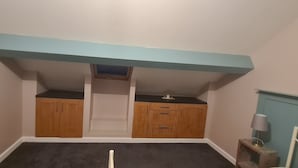 A room with a sloped ceiling, wooden cabinets, a small window, and a lamp on a side table. - Charming 1-bed Apartment in Barnsley (Barnsley)