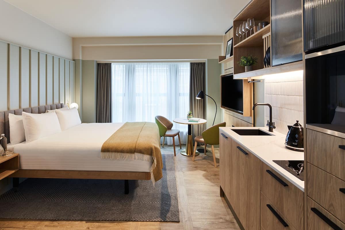 Une chambre d’hôtel comprenant un lit, un petit coin repas, une kitchenette et une grande fenêtre avec des rideaux.