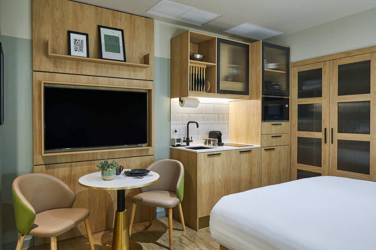 Une chambre d’hôtel compacte comprenant un lit, une petite table à manger avec des chaises, un coin cuisine et un téléviseur fixé au mur.