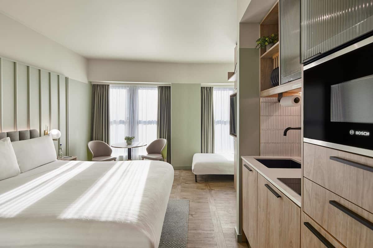 Une chambre d’hôtel avec un grand lit, une petite table avec deux chaises, une kitchenette équipée d’un réfrigérateur Bosch et un téléviseur.