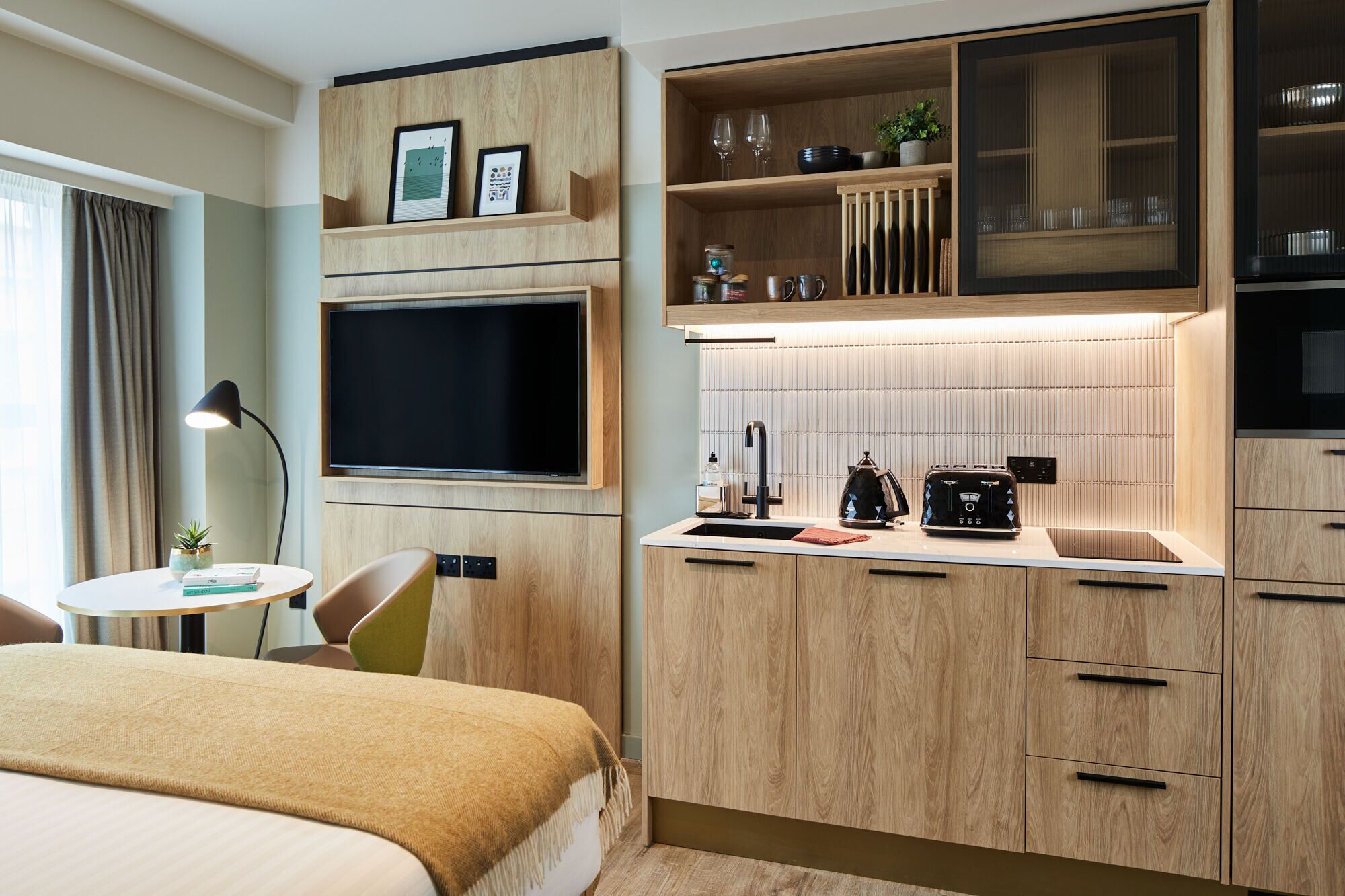 Habitació d'hotel petita amb llit, tauleta, cuineta i televisió penjada a la paret.