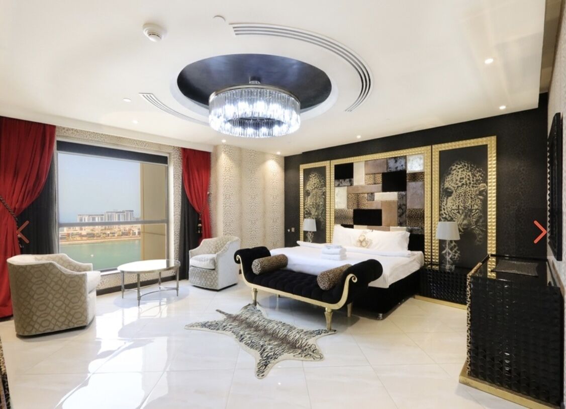 A modern hotel room with a large bed, a chandelier, a city view, and a leather rug.
