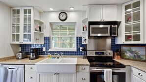 Una cocina con armarios blancos, un microondas de acero inoxidable, un lavamanos doble y una ventana con paredes de azulejos azules.