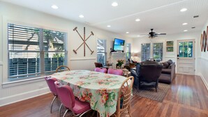 A dining area with a table covered in a floral tablecloth, surrounded by pink chairs. There is a television mounted on the wall and a ceiling fan. - 450 ft to East Beach • 2 Kings • Pet Friendly • Sleeps 6 • St. Simons Island (Saint Simons Island)