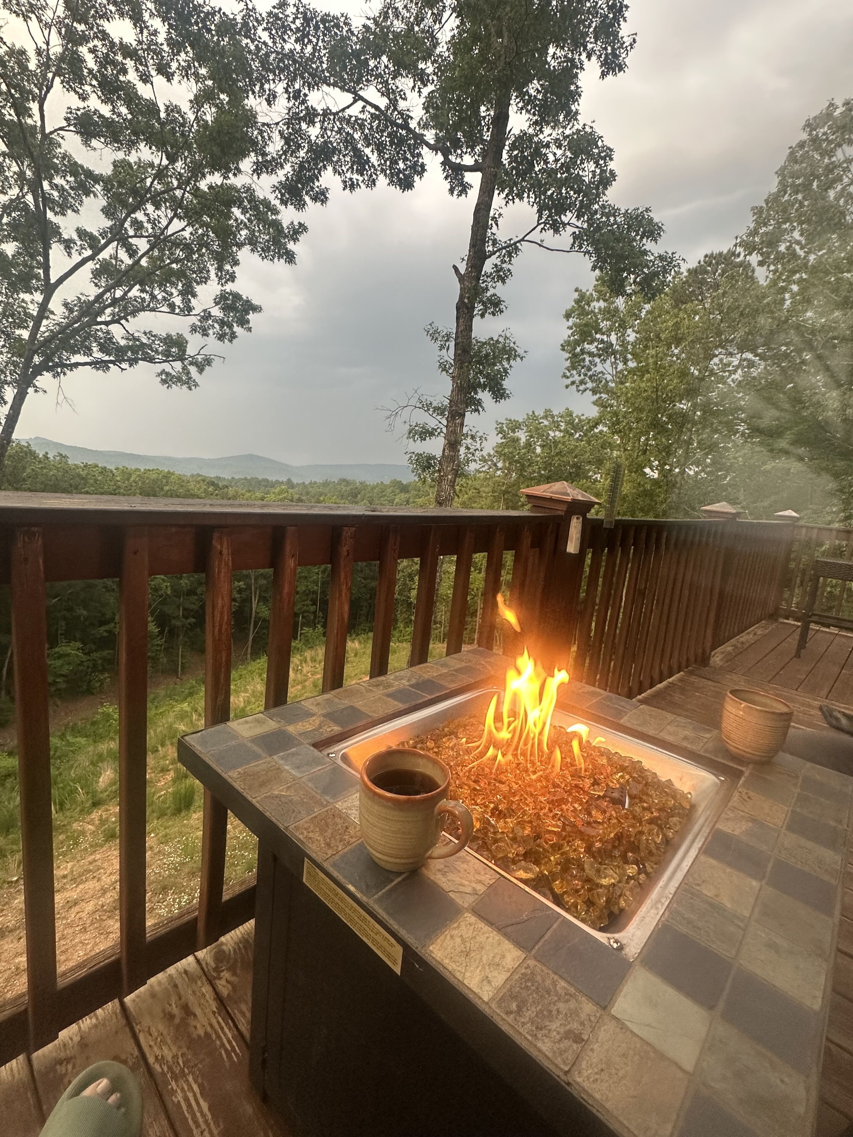 En pejs på en terrasse med udsigt over skov og bjerge i baggrunden.