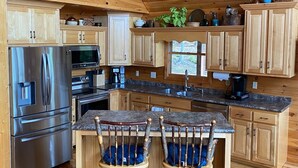 A spacious kitchen with wooden cabinets, a stainless steel refrigerator, and a wooden dining area with two stools. - Mountain Home with Views for Miles!!! (Murphy)