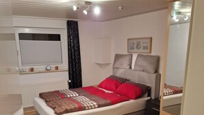 A modern bedroom with a bed, a dresser, a window with blinds, a radiator, and a ceiling with recessed lighting. - Gemütliche Ferienwohnung im Grünen, in der Nähe von Lüneburg (Melbeck)