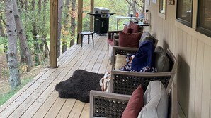 A covered porch with wooden flooring, a seating area with cushions, a grill, and a view of trees.