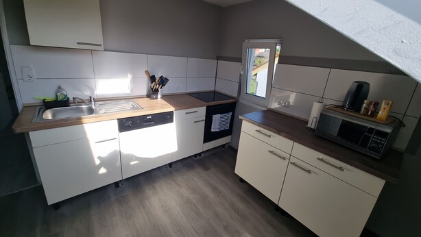 A compact kitchen with white cabinets, a dishwasher, a microwave, and a window providing natural light. - Monteurzimmer Gelnhausen, Ferienwohnung, Auch für Große Gruppen, Nähe Frankfurt (Gelnhausen)