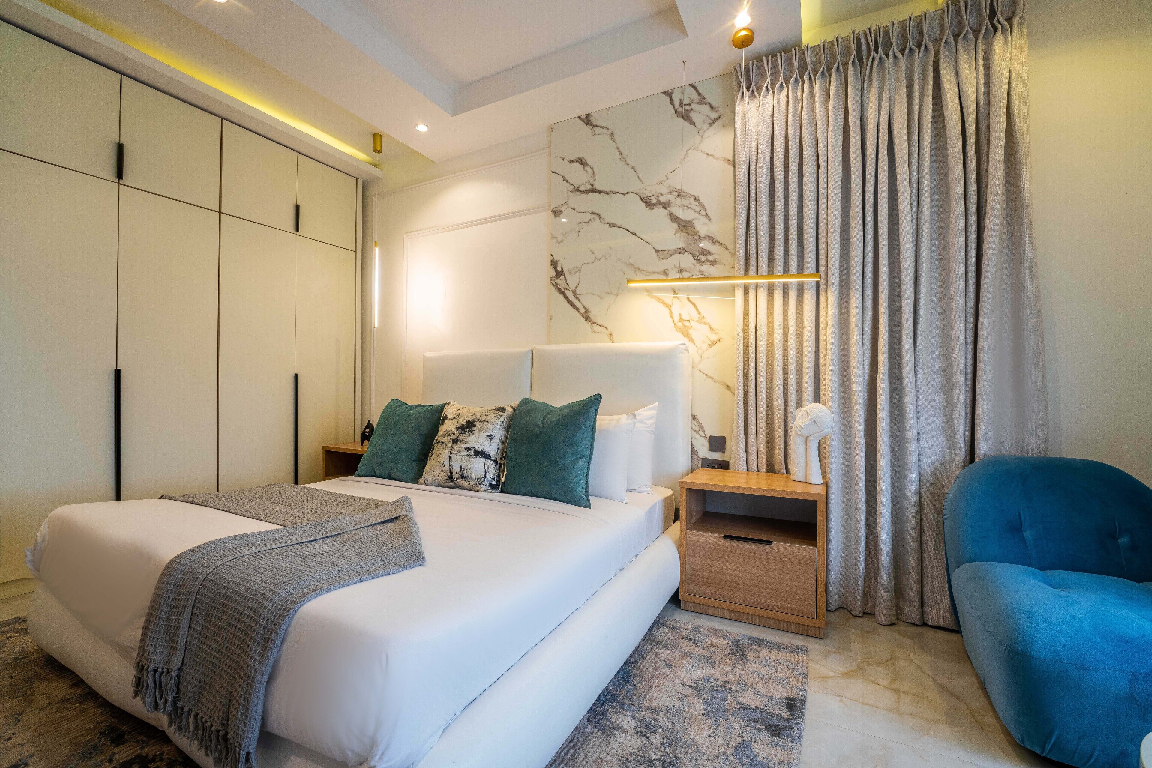 A modern bedroom with a large bed, a nightstand, a blue chair, and a wardrobe.