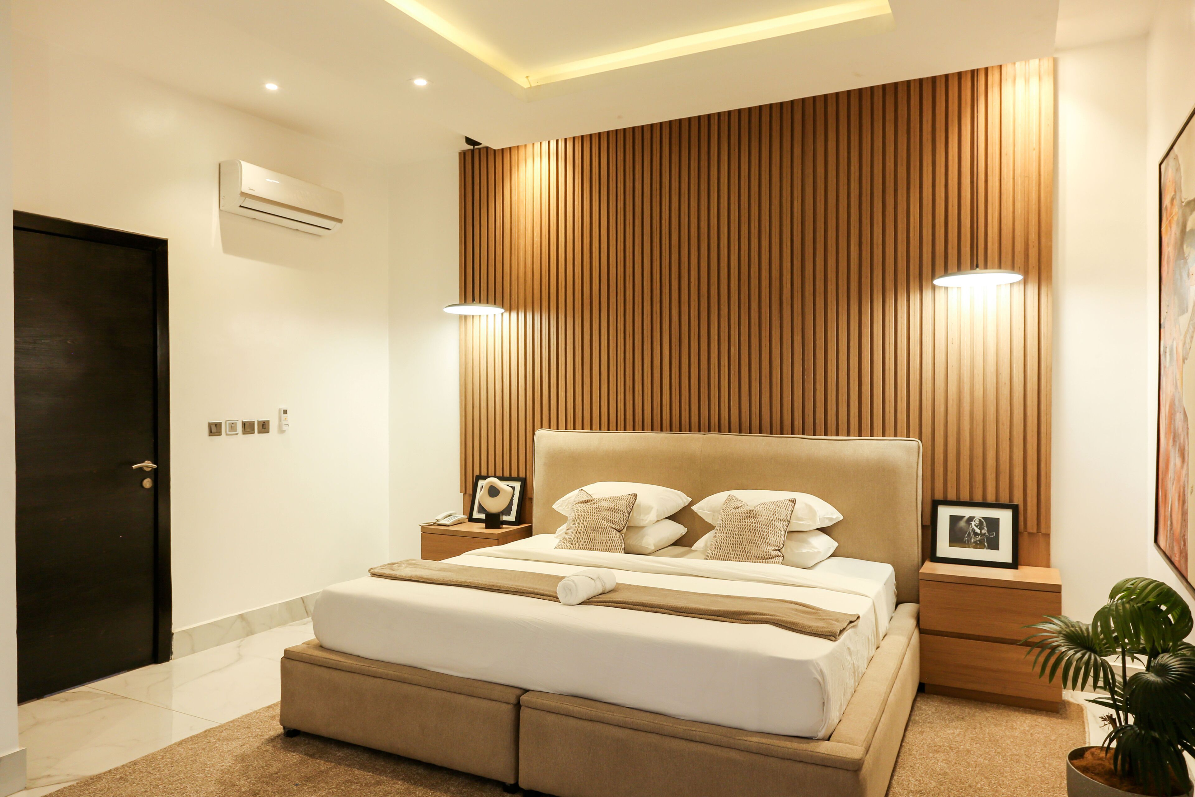 A modern bedroom with a large bed, bedside tables with lamps, a wooden panel wall, and an air conditioning unit.