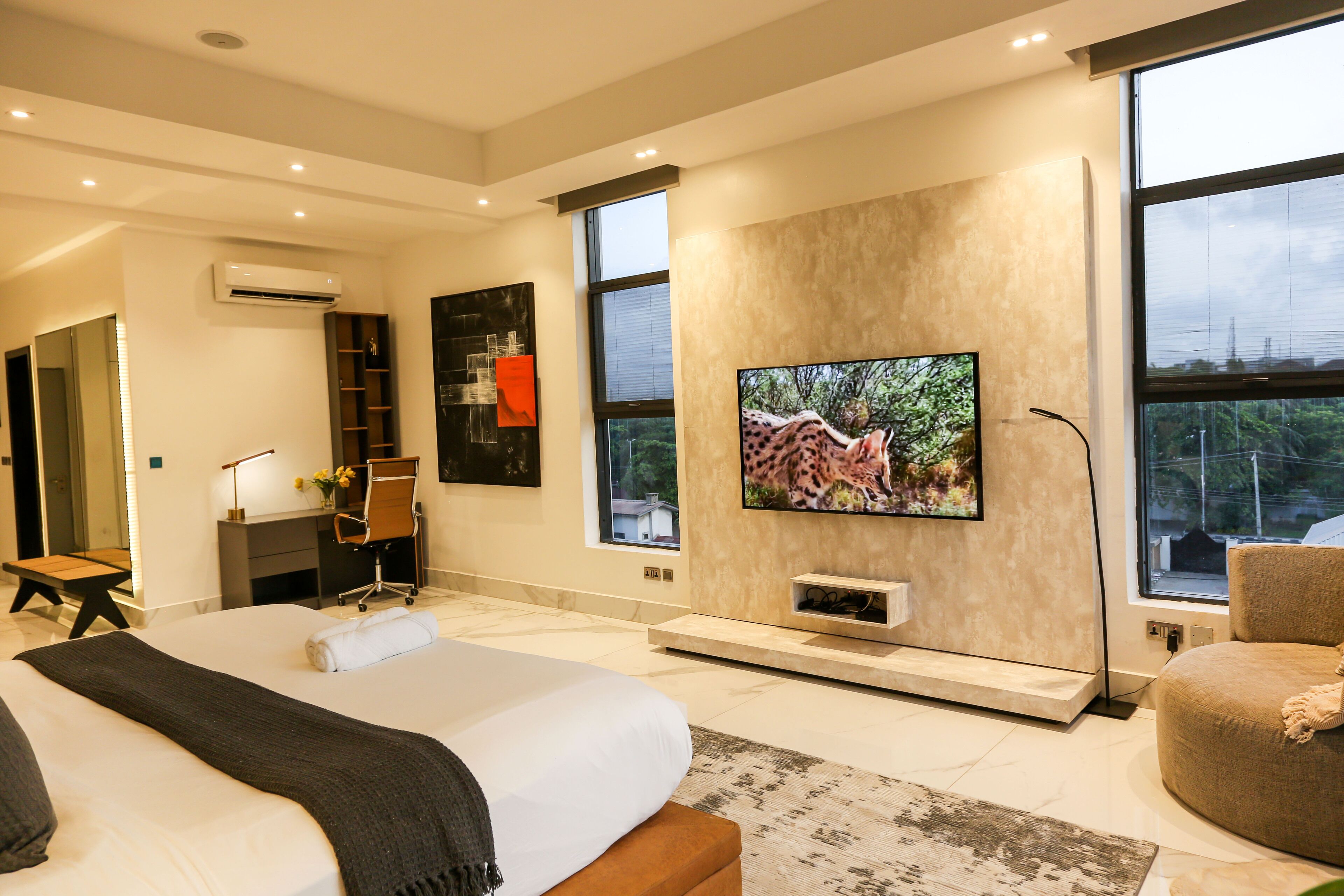 A modern hotel room with a large bed, a desk, a television mounted on the wall, and a large window offering a city view.