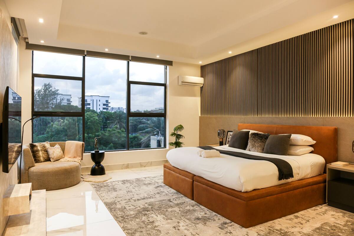 A modern bedroom with a large window, a bed with multiple pillows, a side table, and a wall-mounted air conditioner.