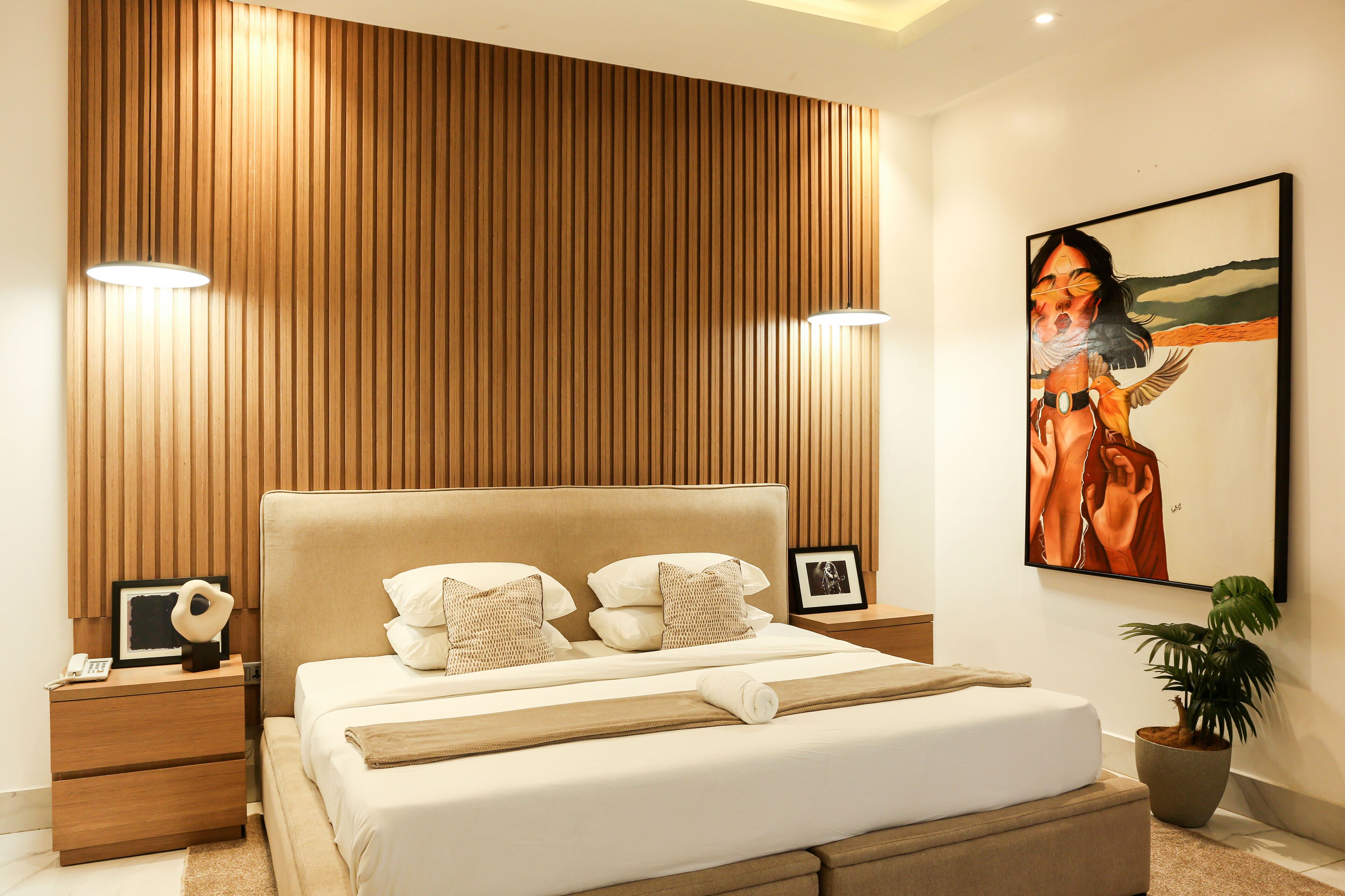 A modern bedroom with a wooden panel wall, a large bed with white bedding, two bedside lamps, a nightstand with a phone, a framed picture, and a potted plant.
