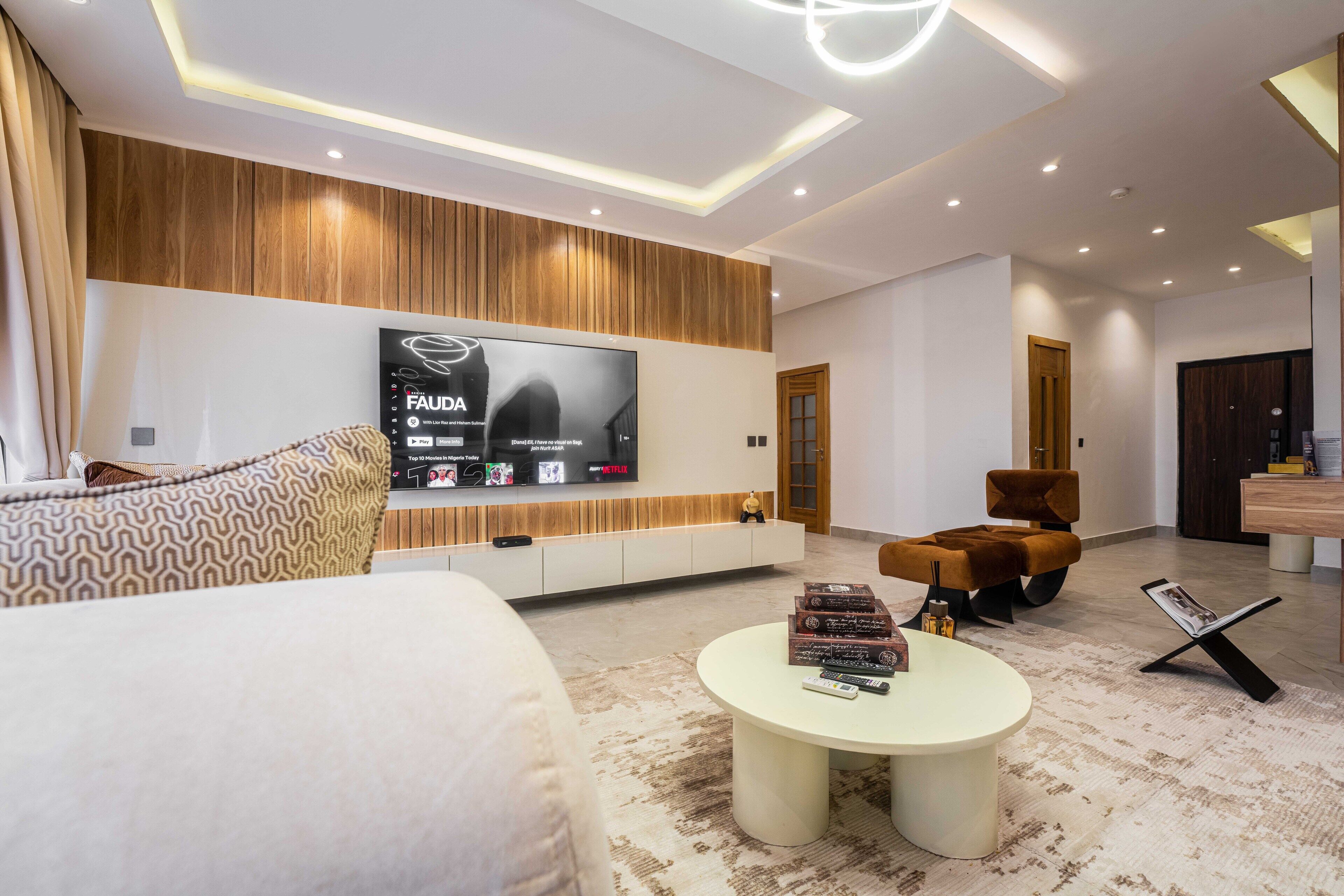 A modern living room with a large flat-screen TV mounted on a wooden wall, a comfortable sofa, a round coffee table with books and remote controls, and a stylish brown armchair.