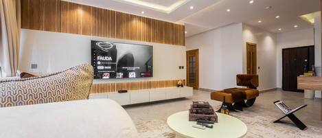 A modern living room with a large flat-screen TV mounted on a wooden wall, a comfortable sofa, a round coffee table with books and remote controls, and a stylish brown armchair.
