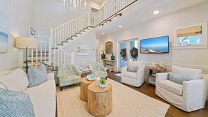A spacious living room with a staircase, white furniture, and a large TV. - Renovated in Seaside, Expansive Porches, 2 Kings, walk to Beach + 2 Bikes! (Santa Rosa Beach)