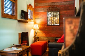 Ein gemütliches Zimmer mit Holzwänden, einem roten Hocker, einem schwarzen Leder Sofa und einem Holztisch sowie einer Lampe.