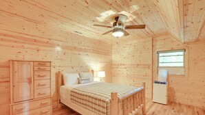 Dormitorio con paneles de madera, una cama, un armario, un ventilador de techo y una ventana.