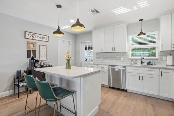 A modern kitchen with white cabinetry, a central island with bar stools, pendant lighting, and a coffee bar sign.