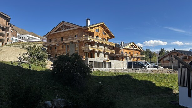 A large wooden chalet with multiple balconies and a stone foundation, surrounded by a grassy area and other buildings. - T2-6pers-34m2-quartier Bergers-pied des Pistes (Huez)